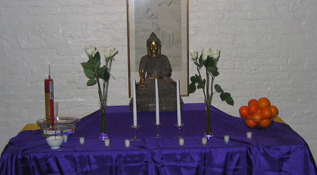 a Tibetan statue on a table with candles, flowers, incense, and oranges